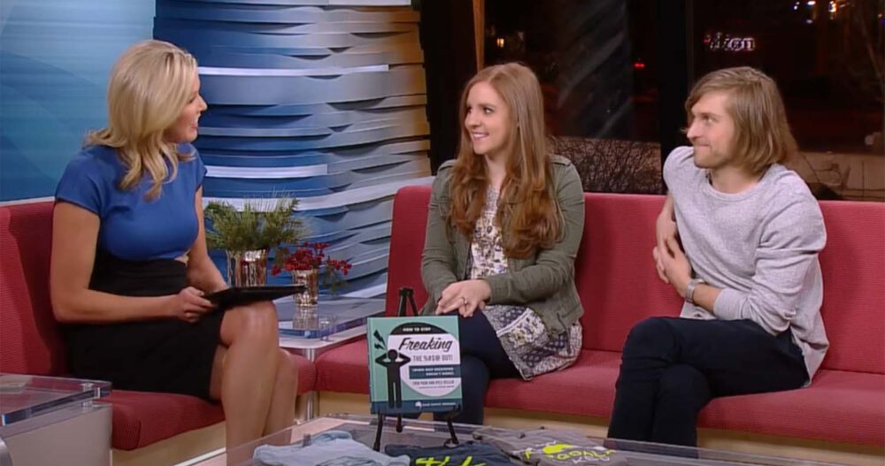 Ellie Mental Health founders Erin Pash and Kyle Keller sitting on a red couch being interviewed by a woman with blond hair wearing a black shirt and blue top about Erin Pash's book, How to Stop Freaking the %#$! Out! (For Couples)