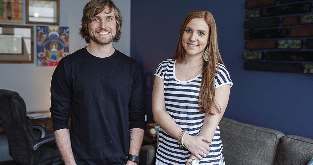 Ellie Mental Health founders Erin Pash, dressed in a white and blue striped shirt, and Kyle Keller, dressed in a dark blue shirt, standing next to each other inside one of the Ellie Mental Health offices.