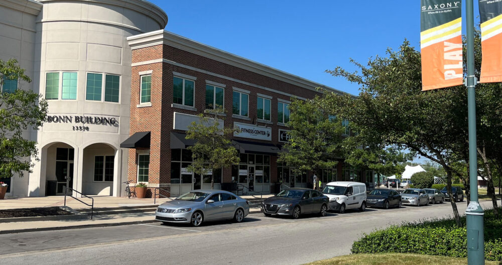 An exterior image of the Ellie Mental Health Fishers, IN building taken from across the street where cars are parked along the street with a bed of bright red flowers and a stylish lamppost in the foreground.