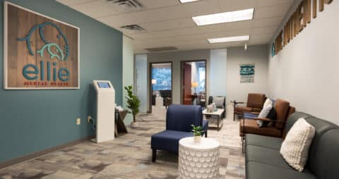 An interior photo of an Ellie Mental Health Therapy Clinic lobby with several couches, chairs, small tables, a check in kiosk, and an Ellie Mental Health logo hanging on one wall.