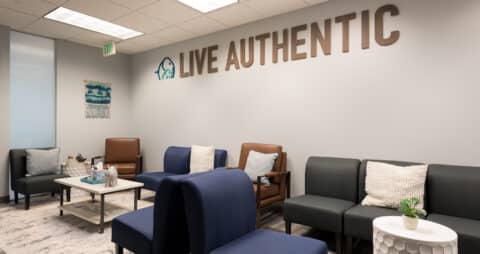 An interior photo of an Ellie Mental Health Therapy Clinic lobby with several blue, gray, and brown chair, small white tables, and "LIVE AUTHENTIC" across one wall next to the Ellie Mental Health logo.