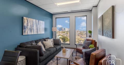 An interior photo of an Ellie Mental Health Therapy Clinic office with two large windows, a dark gray couch, a glass table, two brown leather chairs, and several abstract paintings hanging on the walls.