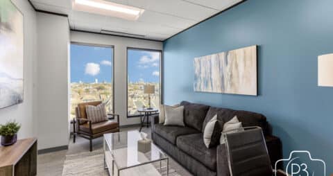 An interior photo of an Ellie Mental Health Therapy Clinic office with two large windows, a gray couch, a glass table, a brown leather chair, and several abstract paintings hanging on the walls.