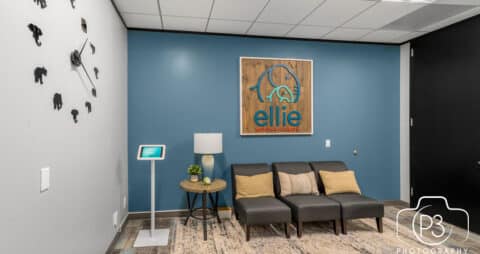 An interior photo of an Ellie Mental Health Therapy Clinic lobby with three dark gray chairs, a small table with plant and table lamp, a check-in kiosk, an Ellie Mental Health logo on one wall and a large clock on another wall.