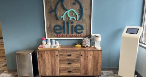 An interior photo of an Ellie Mental Health Therapy Clinic lobby with kiosk check in, cups and coffee supplies sitting on a small wooden desk, and the Ellie Mental Health logo hanging on one wall.