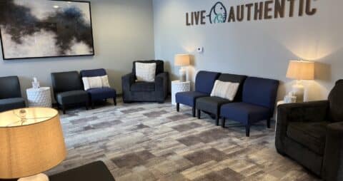 An interior photo of an Ellie Mental Health Therapy Clinic lobby that has stylish gray carpet, blue couches, and the message "LIVE AUTHENTIC" on one wall with the Ellie Mental Health logo.