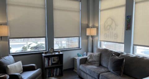 An interior photo of an Ellie Mental Health Therapy Clinic office with four windows, a dark gray couch, a brown leather chair, a small bookcase and two floor lamps.