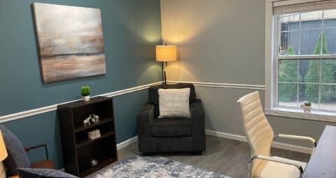 An interior photo of an Ellie Mental Health Therapy Clinic office with a gray chair, floor lamp, a white office chair, a small bookcase, and an abstract painting hanging on the wall.