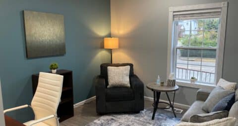 An interior photo of an Ellie Mental Health Therapy Clinic office with a small window, gray couch, dark blue chair, small wooden bookcase, office desk and white office chair, and an abstract painting hanging on one wall.