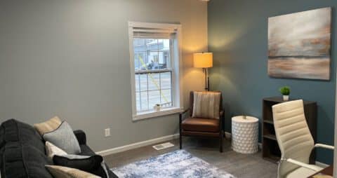 An interior photo of an Ellie Mental Health Therapy Clinic office with a dark blue couch, brown leather chair, small window, floor lamp, small wooden bookcase, white office chair, and an abstract painting hanging on one wall.