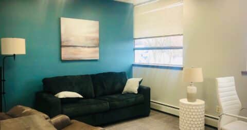 An interior photo of an Ellie Mental Health Therapy Clinic office with dark blue sofa, white office chair, small white table and table lamp, floor lamp and an abstract painting hanging on one wall.