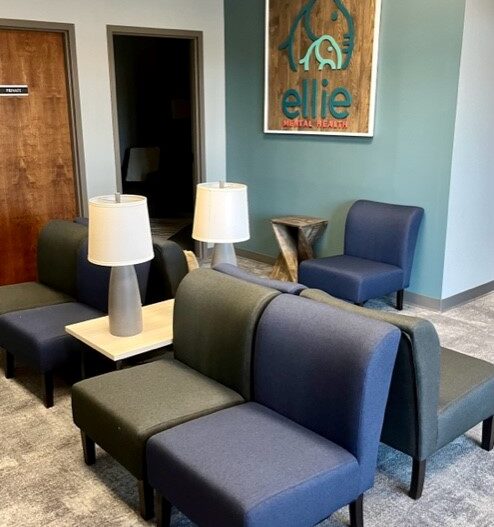 An interior photo of an Ellie Mental Health Therapy Clinic lobby with dark gray and blue chairs, a small table with two gray lamps, and the Ellie Mental Health logo hanging on one wall.
