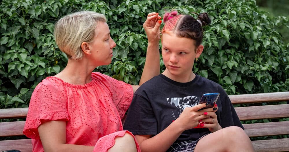 A woman in a pink dress sitting on a bench reaching out to adjust the dark brown and pink hair of a younger woman sitting next to her who's on her phone and has red and black painted fingernails.