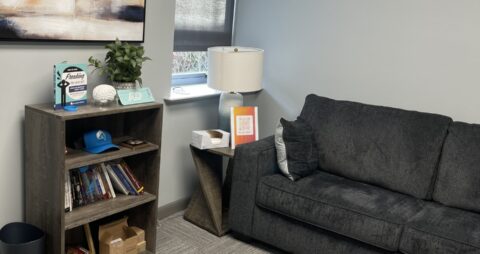 An interior photo of an Ellie Mental Health Therapy Clinic office with dark gray couch, small end table with table lamp, a bookcase with books, and an abstract painting hanging on the wall.