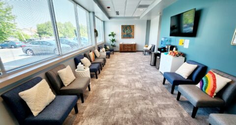 An interior photo of an Ellie Mental Health Therapy Clinic lobby with several blue and gray chairs, a large television on one wall and a painting hanging above a small desk at the far end of the room.
